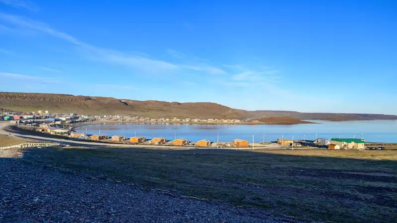Ulukhaktok seen from the bluff: a long shoreline of houses beside calm blue water.